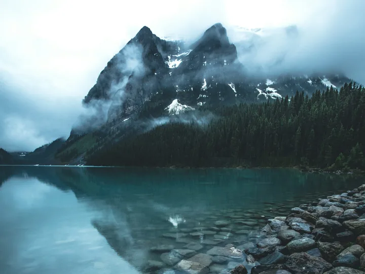 Beispielfoto: Blaugrüner Bergsee mit bewaldetem Ufer vor teils schneebedeckten Felsgipfeln in tiefen Wolken.